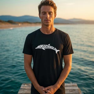 Może przedstawiać: Czarny t-shirt z kr&oacute;tkim rękawem z białą grafiką przedstawiającą sylwetkę delfina zintegrowaną z napisem "BREATHHOLD" na klatce piersiowej. Odzież posiada okrągły dekolt i standardowy kr&oacute;j, wykonana z jednolitego czarnego materiału.