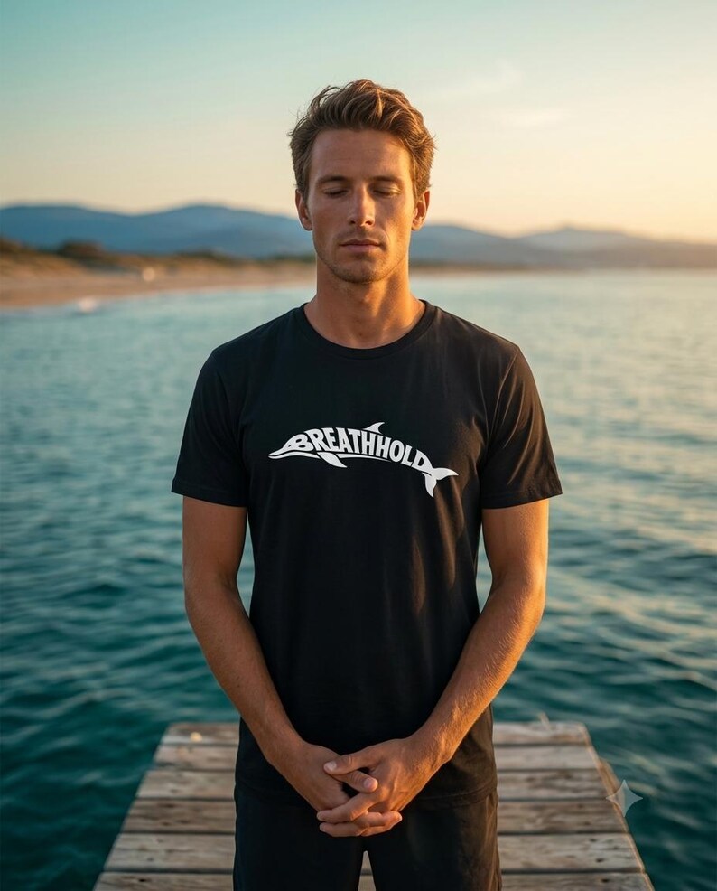 Może przedstawiać: Czarny t-shirt z kr&oacute;tkim rękawem z białą grafiką przedstawiającą sylwetkę delfina zintegrowaną z napisem "BREATHHOLD" na klatce piersiowej. Odzież posiada okrągły dekolt i standardowy kr&oacute;j, wykonana z jednolitego czarnego materiału.