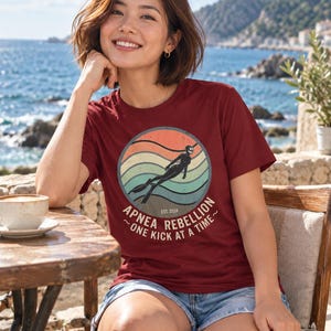 K&ouml;nnte beinhalten: Ein bordeauxrotes Kurzarm-T-Shirt mit einer kreisf&ouml;rmigen Grafik, die einen Retro-Sonnenuntergang, die Silhouette eines Apnoetauchers und den Text "APNEA REBELLION EST. 2024 ~ ONE KICK AT A TIME ~" in heller Schrift auf der Vorderseite zeigt.