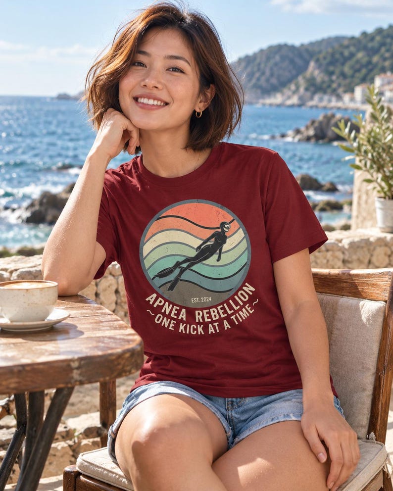 K&ouml;nnte beinhalten: Ein bordeauxrotes Kurzarm-T-Shirt mit einer kreisf&ouml;rmigen Grafik, die einen Retro-Sonnenuntergang, die Silhouette eines Apnoetauchers und den Text "APNEA REBELLION EST. 2024 ~ ONE KICK AT A TIME ~" in heller Schrift auf der Vorderseite zeigt.