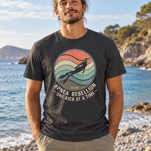 K&ouml;nnte beinhalten: Ein anthrazitfarbenes Kurzarm-T-Shirt mit einer kreisf&ouml;rmigen Grafik im Retro-Sonnenuntergangs-Design in Orange, Beige und T&uuml;rkis. Die Grafik zeigt einen Apnoetaucher sowie den Text "APNEA REBELLION", "ONE KICK AT A TIME" und "EST. 2024".