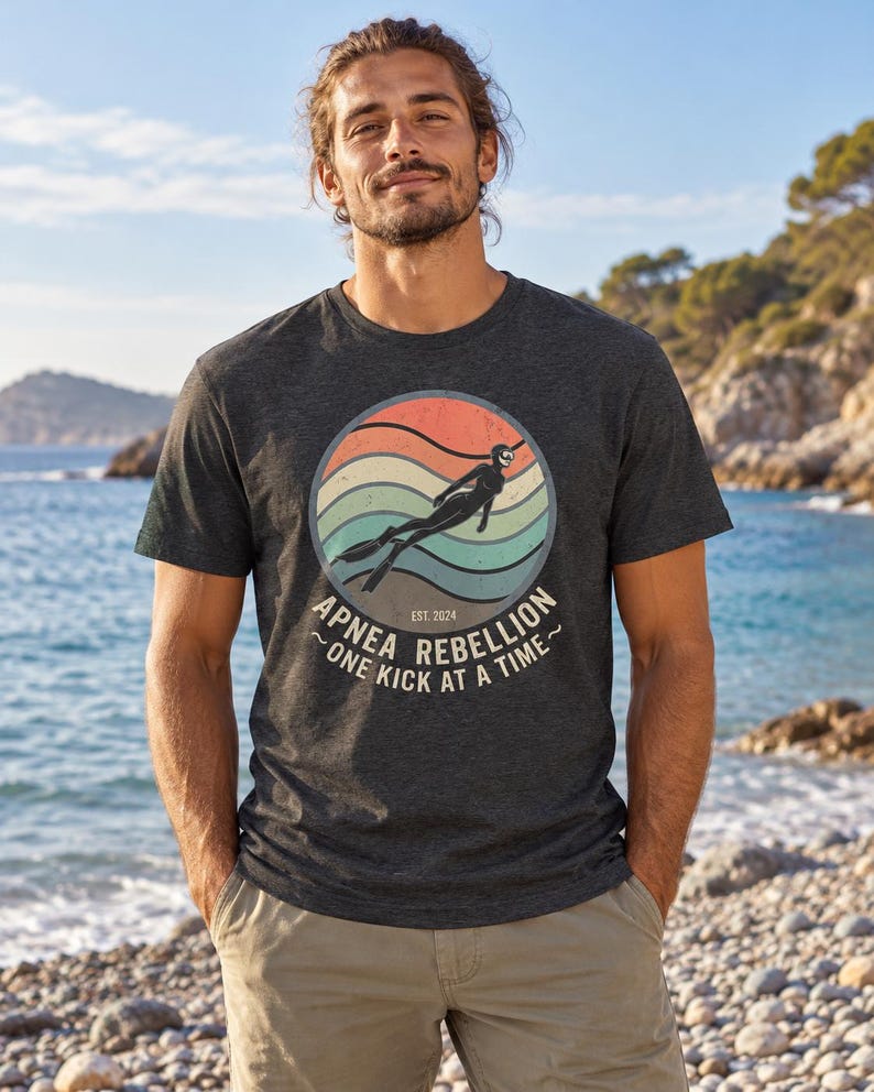 K&ouml;nnte beinhalten: Ein anthrazitfarbenes Kurzarm-T-Shirt mit einer kreisf&ouml;rmigen Grafik im Retro-Sonnenuntergangs-Design in Orange, Beige und T&uuml;rkis. Die Grafik zeigt einen Apnoetaucher sowie den Text "APNEA REBELLION", "ONE KICK AT A TIME" und "EST. 2024".