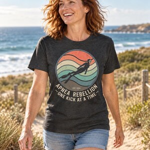 K&ouml;nnte beinhalten: Ein anthrazitfarbenes Kurzarm-T-Shirt mit einem kreisf&ouml;rmigen Aufdruck, der ein Retro-Sonnenuntergangsdesign, die Silhouette eines Apnoetauchers und den Text "APNEA REBELLION EST. 2024 ONE KICK AT A TIME" in verwaschener wei&szlig;er Schrift zeigt.