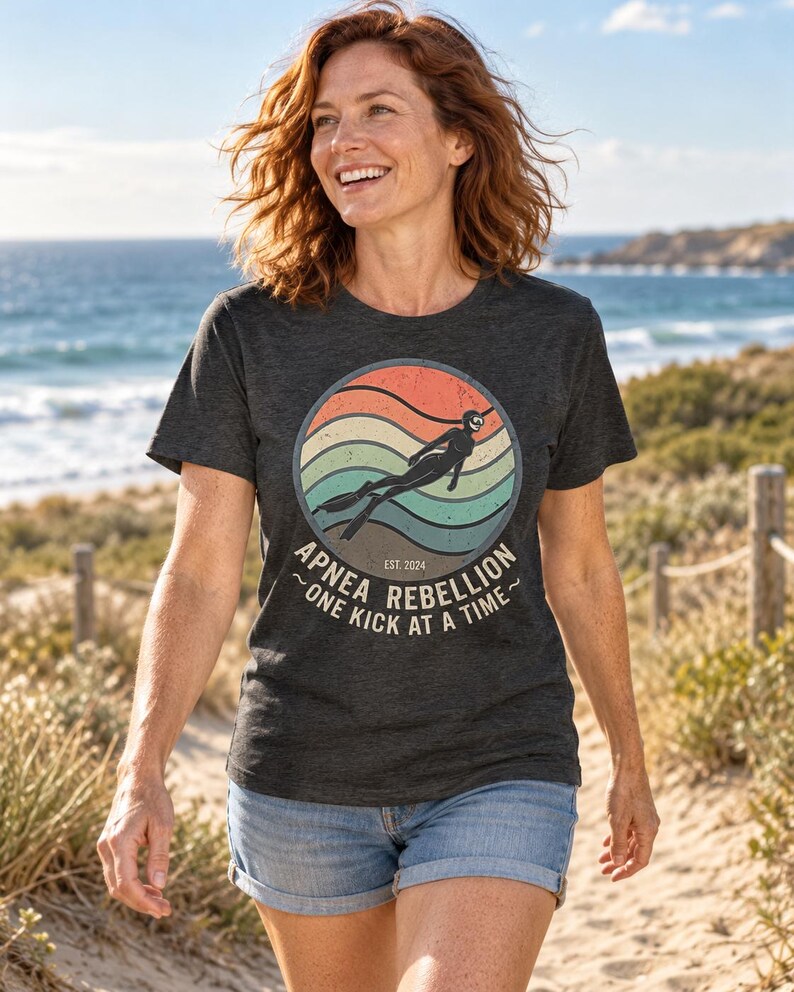K&ouml;nnte beinhalten: Ein anthrazitfarbenes Kurzarm-T-Shirt mit einem kreisf&ouml;rmigen Aufdruck, der ein Retro-Sonnenuntergangsdesign, die Silhouette eines Apnoetauchers und den Text "APNEA REBELLION EST. 2024 ONE KICK AT A TIME" in verwaschener wei&szlig;er Schrift zeigt.