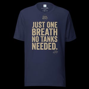 May include: A navy blue short-sleeve t-shirt featuring the beige text JUST ONE BREATH NO TANKS NEEDED. Above the text is a circular logo with the words Apnea Division, and below the text is a smaller circular logo with the words Est 2024 Pure Freedom.