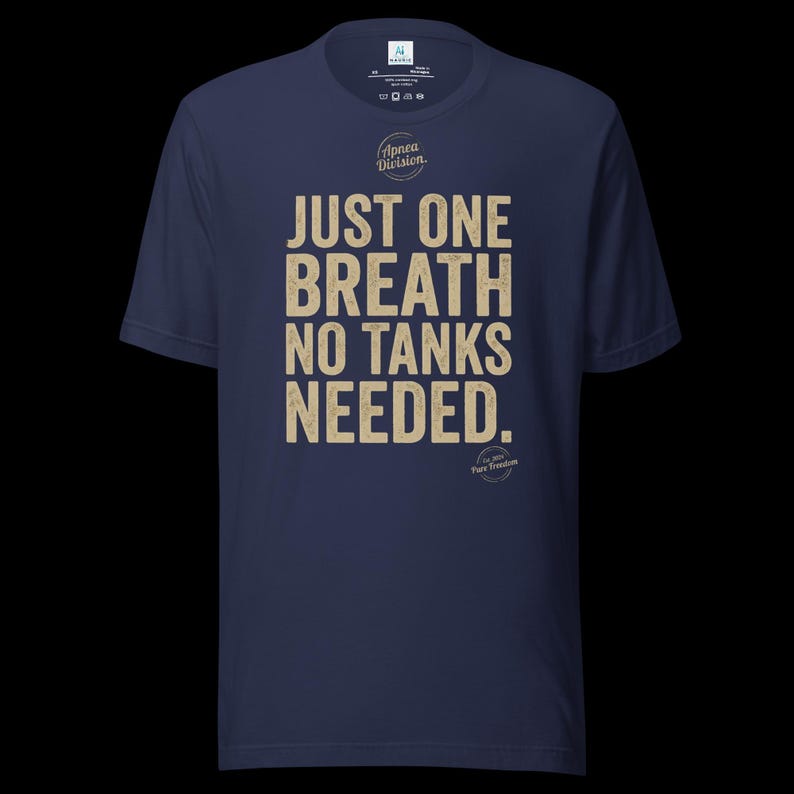 May include: A navy blue short-sleeve t-shirt featuring the beige text JUST ONE BREATH NO TANKS NEEDED. Above the text is a circular logo with the words Apnea Division, and below the text is a smaller circular logo with the words Est 2024 Pure Freedom.