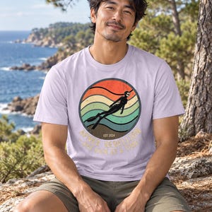 K&ouml;nnte beinhalten: Ein hellviolettes Kurzarm-T-Shirt mit einer kreisf&ouml;rmigen Grafik eines Tauchers im Neoprenanzug vor einem sonnenuntergangsfarbenen Hintergrund mit Wellenlinien. Der Text lautet "APNEA REBELLION" und "ONE KICK AT A TIME" mit "EST. 2024".