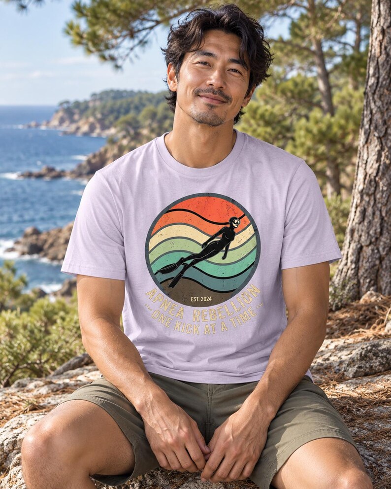 K&ouml;nnte beinhalten: Ein hellviolettes Kurzarm-T-Shirt mit einer kreisf&ouml;rmigen Grafik eines Tauchers im Neoprenanzug vor einem sonnenuntergangsfarbenen Hintergrund mit Wellenlinien. Der Text lautet "APNEA REBELLION" und "ONE KICK AT A TIME" mit "EST. 2024".