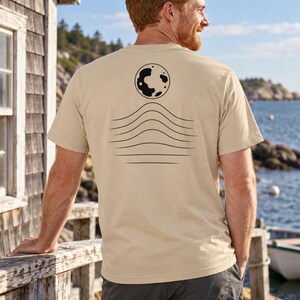 K&ouml;nnte beinhalten: Ein beiges T-Shirt mit kurzen &Auml;rmeln und einer schwarzen Grafik auf der R&uuml;ckseite. Das Design besteht aus einer kreisf&ouml;rmigen Mondform &uuml;ber einer Reihe von f&uuml;nf horizontalen, wellenf&ouml;rmigen Linien, die sich in der Mitte nach oben w&ouml;lben.