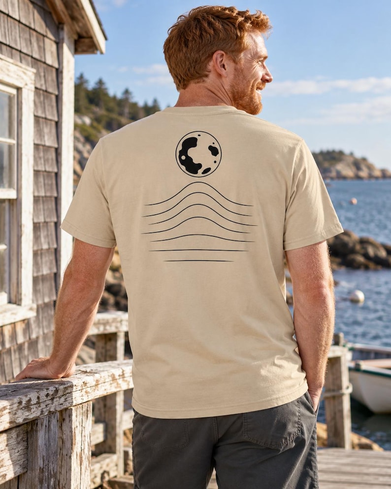 K&ouml;nnte beinhalten: Ein beiges T-Shirt mit kurzen &Auml;rmeln und einer schwarzen Grafik auf der R&uuml;ckseite. Das Design besteht aus einer kreisf&ouml;rmigen Mondform &uuml;ber einer Reihe von f&uuml;nf horizontalen, wellenf&ouml;rmigen Linien, die sich in der Mitte nach oben w&ouml;lben.