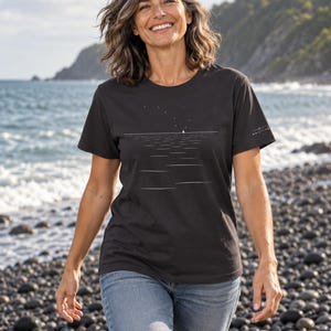 Peut inclure: Un t-shirt noir &agrave; manches courtes orn&eacute; d'un graphisme minimaliste en lignes blanches repr&eacute;sentant un voilier &agrave; l'horizon sous un ciel &eacute;toil&eacute;. Le design inclut un petit logo blanc sur la manche gauche. Le t-shirt est port&eacute; avec un jean bleu.