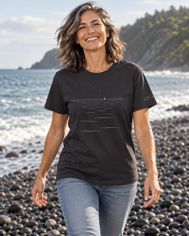 Peut inclure: Un t-shirt noir &agrave; manches courtes orn&eacute; d'un graphisme minimaliste en lignes blanches repr&eacute;sentant un voilier &agrave; l'horizon sous un ciel &eacute;toil&eacute;. Le design inclut un petit logo blanc sur la manche gauche. Le t-shirt est port&eacute; avec un jean bleu.