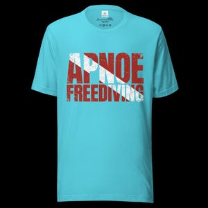 Puede incluir: Una camiseta de manga corta de color turquesa brillante con un estampado gr&aacute;fico desgastado en rojo y blanco. El dise&ntilde;o muestra el texto APNOE FREEDIVING superpuesto sobre un patr&oacute;n de rayas diagonales rojas y blancas, similar a una bandera de buceo.