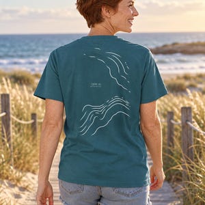 K&ouml;nnte beinhalten: Ein blaugr&uuml;nes Kurzarm-T-Shirt mit einer wei&szlig;en Linien-Grafik auf der R&uuml;ckseite. Das Design zeigt abstrakte Wellenmuster und enth&auml;lt den Text -1200 m ATLANTIC, der in Wei&szlig; zwischen den beiden Wellenillustrationen auf dem R&uuml;cken gedruckt ist.