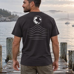 K&ouml;nnte beinhalten: Ein anthrazitfarbenes Kurzarm-T-Shirt mit einer wei&szlig;en Grafik auf der R&uuml;ckseite. Das Design zeigt einen kreisf&ouml;rmigen Mond &uuml;ber einer Reihe von f&uuml;nf horizontalen, wellenf&ouml;rmigen Linien, die nach unten hin k&uuml;rzer werden.