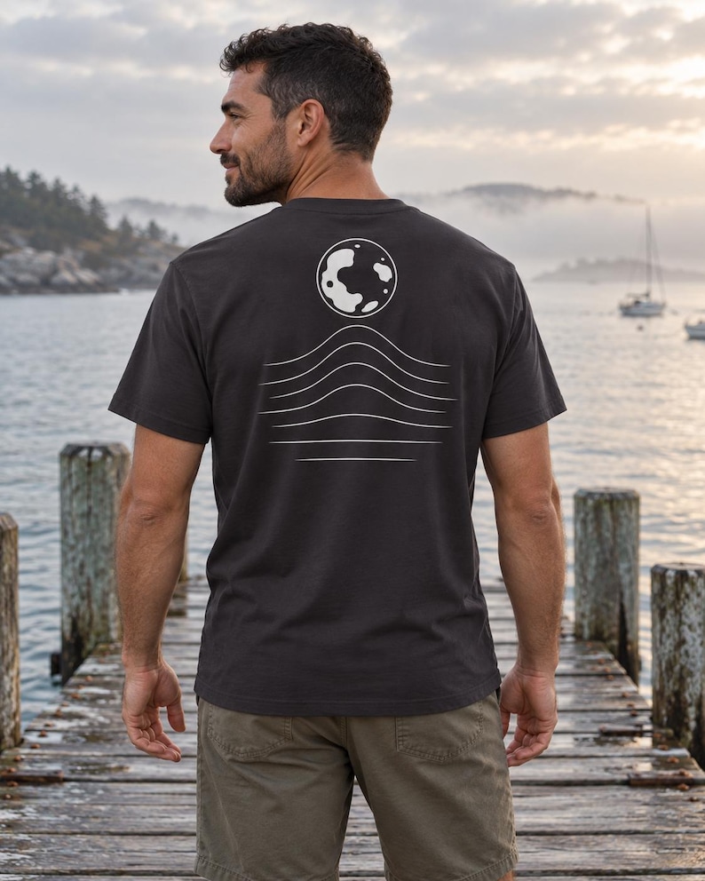 K&ouml;nnte beinhalten: Ein anthrazitfarbenes Kurzarm-T-Shirt mit einer wei&szlig;en Grafik auf der R&uuml;ckseite. Das Design zeigt einen kreisf&ouml;rmigen Mond &uuml;ber einer Reihe von f&uuml;nf horizontalen, wellenf&ouml;rmigen Linien, die nach unten hin k&uuml;rzer werden.