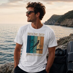 Puede incluir: Una camiseta blanca de manga corta con un estampado gr&aacute;fico que incluye el texto INTO THE DEEP y Apnea Experience. El dise&ntilde;o presenta un fondo de rayas verticales en azul, verde y naranja, con la silueta de un buceador y un tibur&oacute;n.