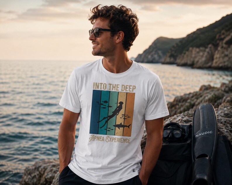 Puede incluir: Una camiseta blanca de manga corta con un estampado gr&aacute;fico que incluye el texto INTO THE DEEP y Apnea Experience. El dise&ntilde;o presenta un fondo de rayas verticales en azul, verde y naranja, con la silueta de un buceador y un tibur&oacute;n.
