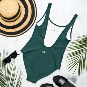 Peut inclure: Un maillot de bain une pi&egrave;ce vert fonc&eacute; avec une doublure int&eacute;rieure blanche portant l'inscription NAUTIVE. L'ext&eacute;rieur pr&eacute;sente un motif de lignes topographiques subtil et un petit logo vague blanc, accompagn&eacute; d'un chapeau de paille et de lunettes.