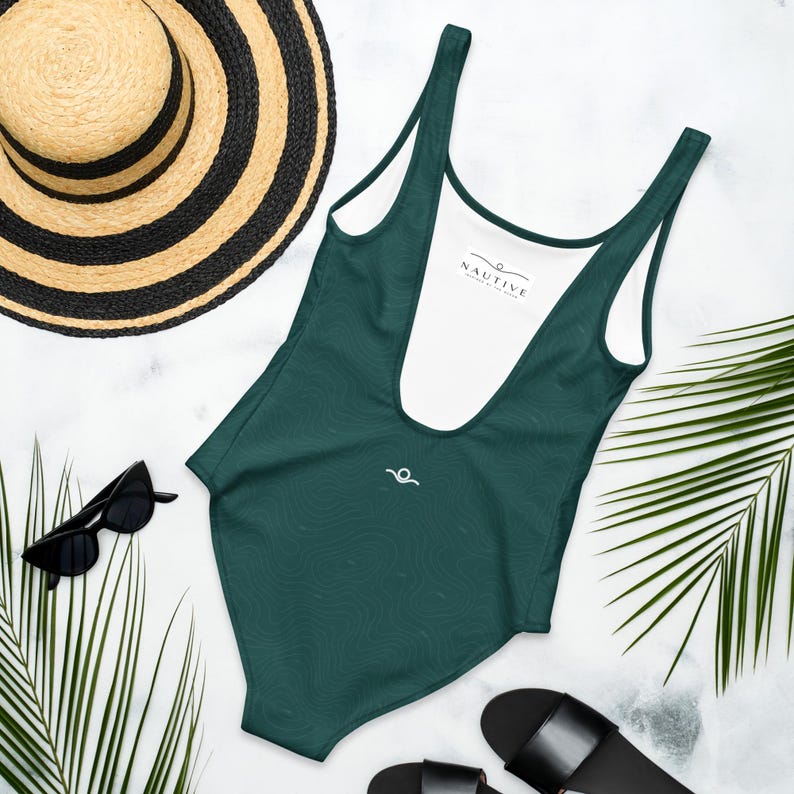 Peut inclure: Un maillot de bain une pi&egrave;ce vert fonc&eacute; avec une doublure int&eacute;rieure blanche portant l'inscription NAUTIVE. L'ext&eacute;rieur pr&eacute;sente un motif de lignes topographiques subtil et un petit logo vague blanc, accompagn&eacute; d'un chapeau de paille et de lunettes.