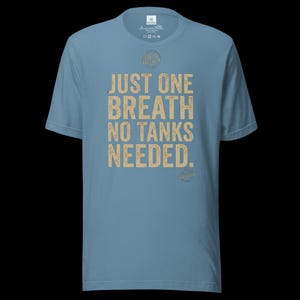 May include: A dusty blue short-sleeve t-shirt featuring the text JUST ONE BREATH NO TANKS NEEDED in a distressed tan font. The shirt includes a small circular graphic above the text and a smaller circular logo at the bottom right of the print.
