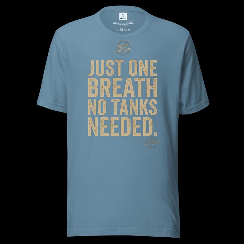 May include: A dusty blue short-sleeve t-shirt featuring the text JUST ONE BREATH NO TANKS NEEDED in a distressed tan font. The shirt includes a small circular graphic above the text and a smaller circular logo at the bottom right of the print.