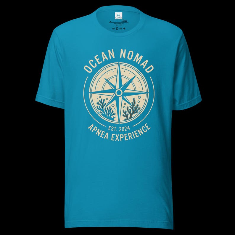 Op de afbeelding: Een felblauw t-shirt met korte mouwen en een cr&egrave;mekleurige cirkelvormige afbeelding op de borst. Het ontwerp bevat een kompasroos, koraalillustraties en de tekst "OCEAN NOMAD" en "APNEA EXPERIENCE EST. 2024" in een strak, schreefloos lettertype.
