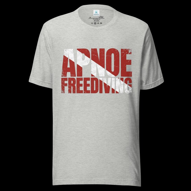 Puede incluir: Una camiseta de manga corta en color gris jaspeado claro con un estampado gr&aacute;fico desgastado en rojo y blanco en el pecho. El dise&ntilde;o muestra el texto APNOE FREEDIVING sobre un patr&oacute;n de rayas diagonales rojas y blancas que recuerda a una bandera de buceo.