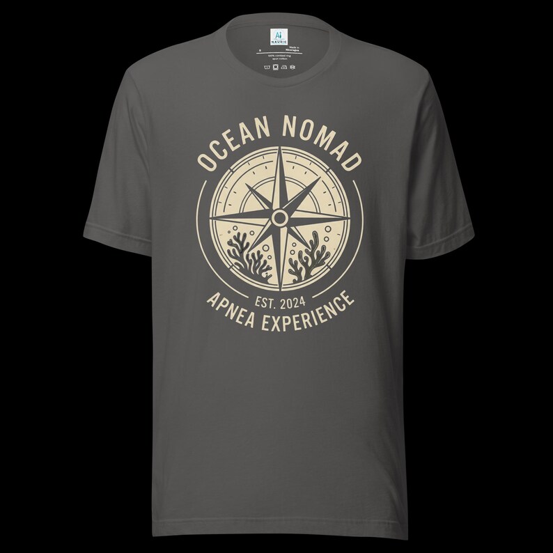 Op de afbeelding: Een antracietgrijs T-shirt met korte mouwen en een cr&egrave;mekleurige cirkelvormige afbeelding op de borst. Het ontwerp bevat een kompasroos, koraalillustraties en de tekst "OCEAN NOMAD" en "APNEA EXPERIENCE EST. 2024" in een cirkelvormige lay-out.