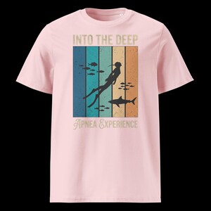 Puede incluir: Una camiseta rosa claro con un estampado gr&aacute;fico que incluye el texto INTO THE DEEP y APNEA EXPERIENCE. El dise&ntilde;o presenta un fondo de rayas verticales en tonos azul, turquesa y naranja, con la silueta de un buceador y un tibur&oacute;n nadando.