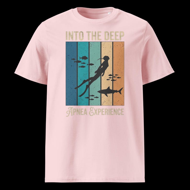 Puede incluir: Una camiseta rosa claro con un estampado gr&aacute;fico que incluye el texto INTO THE DEEP y APNEA EXPERIENCE. El dise&ntilde;o presenta un fondo de rayas verticales en tonos azul, turquesa y naranja, con la silueta de un buceador y un tibur&oacute;n nadando.
