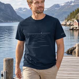 Peut inclure: Un t-shirt bleu marine &agrave; manches courtes orn&eacute; d'un graphisme lin&eacute;aire blanc repr&eacute;sentant un petit voilier sur l'eau sous un ciel &eacute;toil&eacute;. La manche gauche porte un petit logo blanc avec le texte NAUTIV. Le t-shirt a un col rond et un ourlet droit.