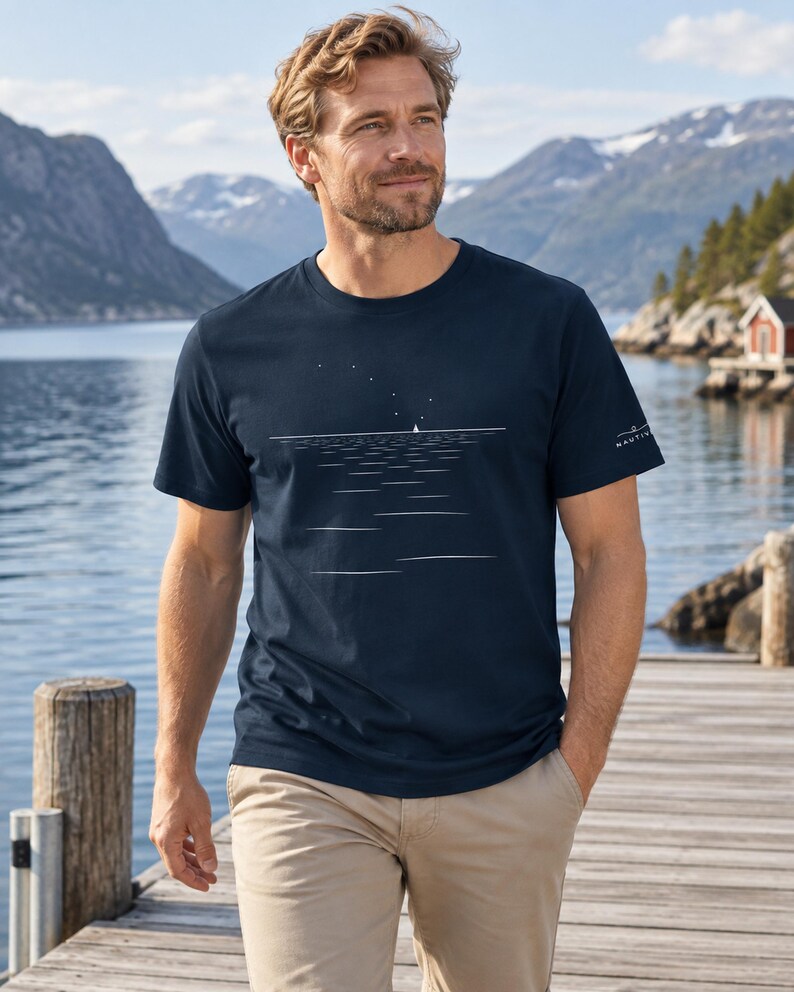 Peut inclure: Un t-shirt bleu marine &agrave; manches courtes orn&eacute; d'un graphisme lin&eacute;aire blanc repr&eacute;sentant un petit voilier sur l'eau sous un ciel &eacute;toil&eacute;. La manche gauche porte un petit logo blanc avec le texte NAUTIV. Le t-shirt a un col rond et un ourlet droit.