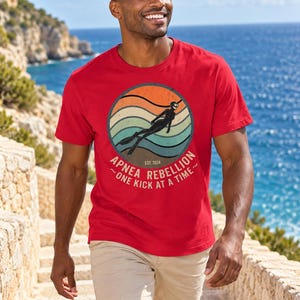 K&ouml;nnte beinhalten: Ein rotes Kurzarm-T-Shirt mit einem kreisf&ouml;rmigen Grafikdruck, der ein Retro-Sonnenuntergangsdesign, die Silhouette eines Apnoetauchers sowie den Text "APNEA REBELLION ~ONE KICK AT A TIME~" und "EST. 2024" in einer Vintage-Schriftart zeigt.