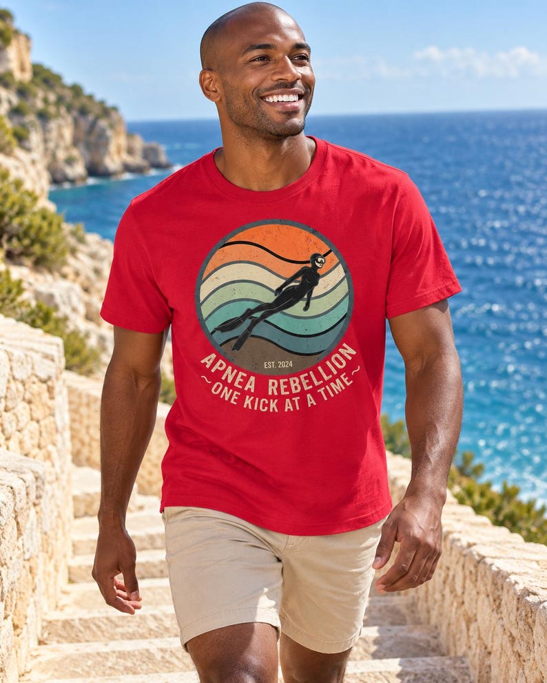 K&ouml;nnte beinhalten: Ein rotes Kurzarm-T-Shirt mit einem kreisf&ouml;rmigen Grafikdruck, der ein Retro-Sonnenuntergangsdesign, die Silhouette eines Apnoetauchers sowie den Text "APNEA REBELLION ~ONE KICK AT A TIME~" und "EST. 2024" in einer Vintage-Schriftart zeigt.