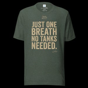 May include: A heathered olive green short-sleeve t-shirt featuring the beige text "JUST ONE BREATH NO TANKS NEEDED." and the "Apnea Division" logo printed on the chest. The shirt has a crew neckline and a standard fit for casual wear.