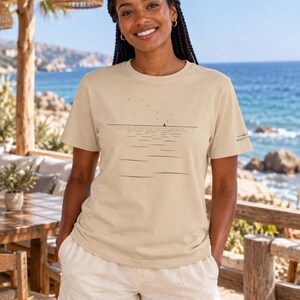 Peut inclure: Un t-shirt beige &agrave; manches courtes orn&eacute; d'un graphique minimaliste en lignes noires repr&eacute;sentant une ligne d'horizon, un petit voilier et des points dispers&eacute;s &eacute;voquant des oiseaux. La manche gauche porte un petit logo noir Nautica.