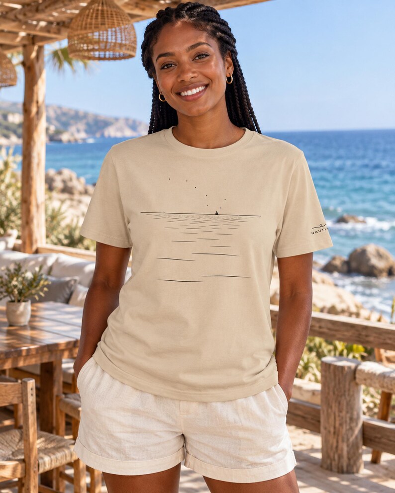 Peut inclure: Un t-shirt beige &agrave; manches courtes orn&eacute; d'un graphique minimaliste en lignes noires repr&eacute;sentant une ligne d'horizon, un petit voilier et des points dispers&eacute;s &eacute;voquant des oiseaux. La manche gauche porte un petit logo noir Nautica.