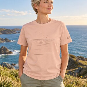 Peut inclure: Un t-shirt &agrave; manches courtes de couleur p&ecirc;che avec un dessin au trait noir minimaliste repr&eacute;sentant une ligne d'horizon, un petit voilier et des oiseaux dans le ciel. La manche gauche porte le texte NAUTIVO en petite police noire.