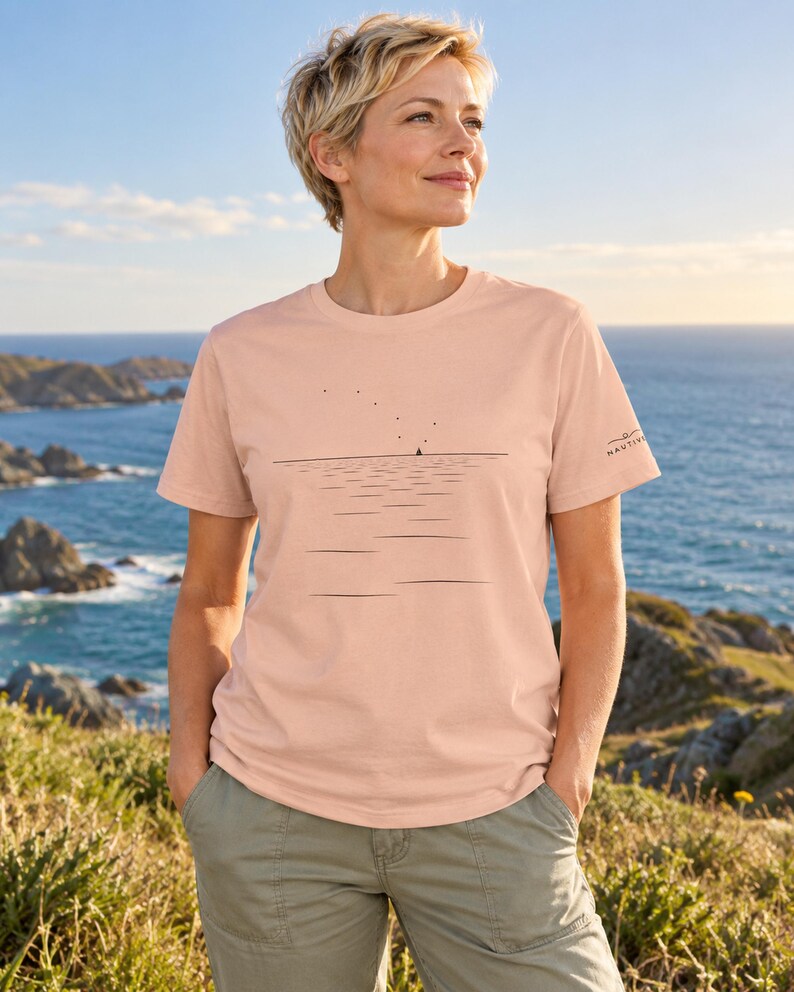Peut inclure: Un t-shirt &agrave; manches courtes de couleur p&ecirc;che avec un dessin au trait noir minimaliste repr&eacute;sentant une ligne d'horizon, un petit voilier et des oiseaux dans le ciel. La manche gauche porte le texte NAUTIVO en petite police noire.