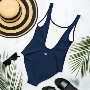Peut inclure: Un maillot de bain une pi&egrave;ce bleu marine fonc&eacute; avec un motif subtil de lignes topographiques, un d&eacute;collet&eacute; plongeant et une &eacute;tiquette int&eacute;rieure blanche portant le texte NAUTIVE. Il pr&eacute;sente un petit logo blanc en forme de vague sur la poitrine.