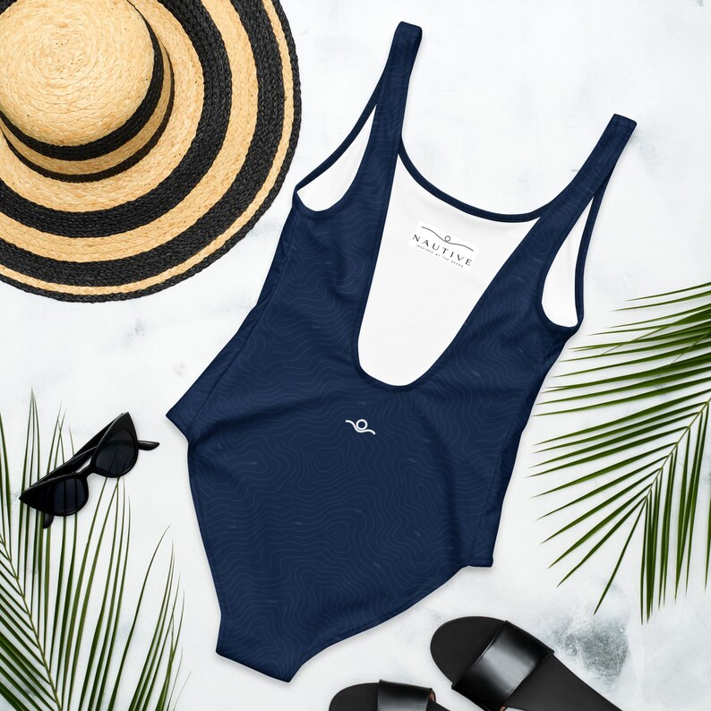 Peut inclure: Un maillot de bain une pi&egrave;ce bleu marine fonc&eacute; avec un motif subtil de lignes topographiques, un d&eacute;collet&eacute; plongeant et une &eacute;tiquette int&eacute;rieure blanche portant le texte NAUTIVE. Il pr&eacute;sente un petit logo blanc en forme de vague sur la poitrine.