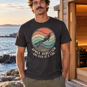 K&ouml;nnte beinhalten: Ein anthrazitfarbenes Kurzarm-T-Shirt mit einer kreisf&ouml;rmigen Grafik auf sonnenuntergangsfarbenem Hintergrund, einer Silhouette eines Apnoetauchers und dem Text "APNEA REBELLION EST. 2024 ~ONE KICK AT A TIME~". Das T-Shirt hat einen Rundhalsausschnitt.
