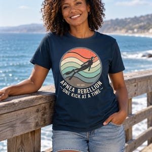 K&ouml;nnte beinhalten: Ein dunkelblaues Kurzarm-T-Shirt mit einer kreisf&ouml;rmigen Grafik, die einen Retro-Sonnenuntergang in Orange, Beige und T&uuml;rkis zeigt. Das Motiv stellt einen Apnoetaucher dar und enth&auml;lt den Text "APNEA REBELLION", "EST. 2024" und "ONE KICK AT A TIME".