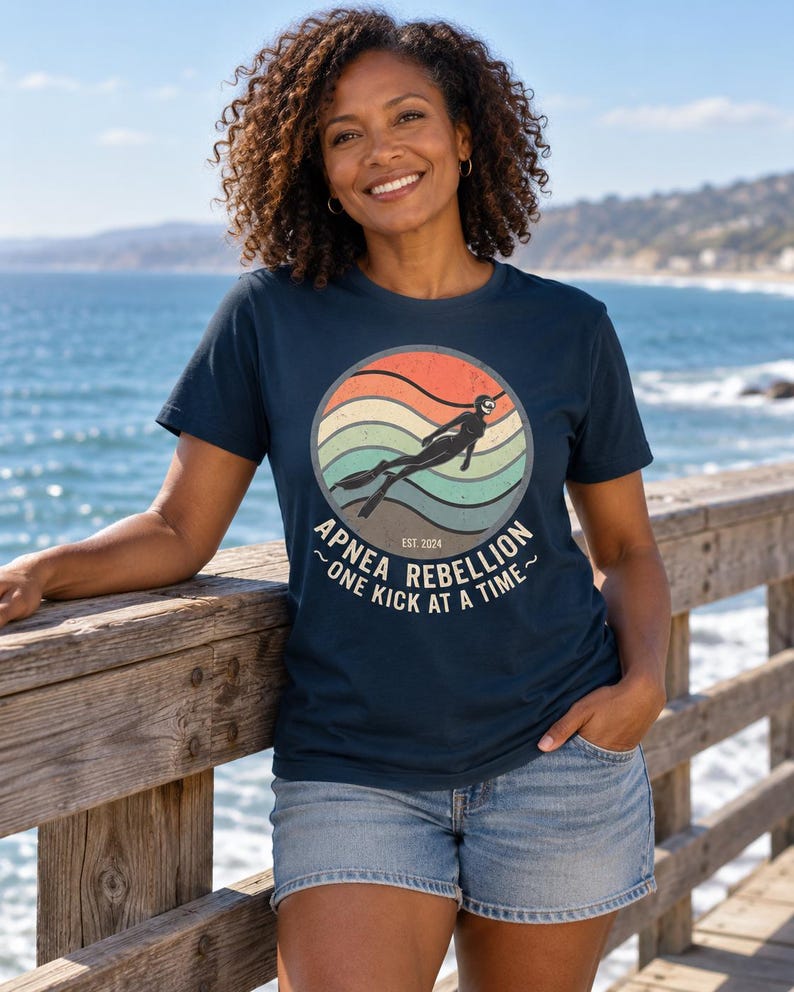 K&ouml;nnte beinhalten: Ein dunkelblaues Kurzarm-T-Shirt mit einer kreisf&ouml;rmigen Grafik, die einen Retro-Sonnenuntergang in Orange, Beige und T&uuml;rkis zeigt. Das Motiv stellt einen Apnoetaucher dar und enth&auml;lt den Text "APNEA REBELLION", "EST. 2024" und "ONE KICK AT A TIME".
