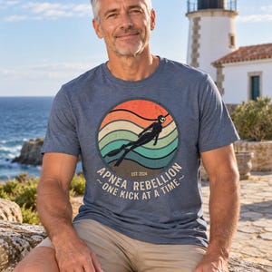K&ouml;nnte beinhalten: Ein kurz&auml;rmeliges T-Shirt in Blau meliert mit einem kreisf&ouml;rmigen Aufdruck, der ein Retro-Sonnenuntergangsdesign, die Silhouette eines Apnoetauchers und den Text "APNEA REBELLION EST. 2024 ~ONE KICK AT A TIME~" zeigt. Es hat einen Rundhalsausschnitt.