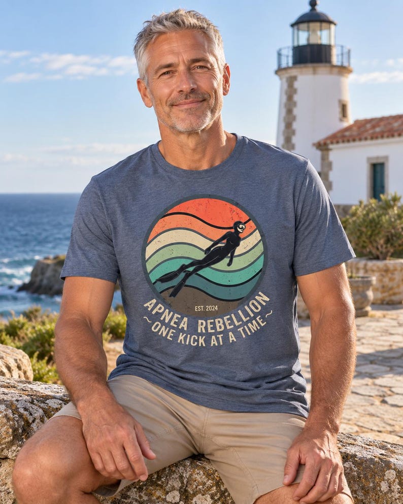 K&ouml;nnte beinhalten: Ein kurz&auml;rmeliges T-Shirt in Blau meliert mit einem kreisf&ouml;rmigen Aufdruck, der ein Retro-Sonnenuntergangsdesign, die Silhouette eines Apnoetauchers und den Text "APNEA REBELLION EST. 2024 ~ONE KICK AT A TIME~" zeigt. Es hat einen Rundhalsausschnitt.