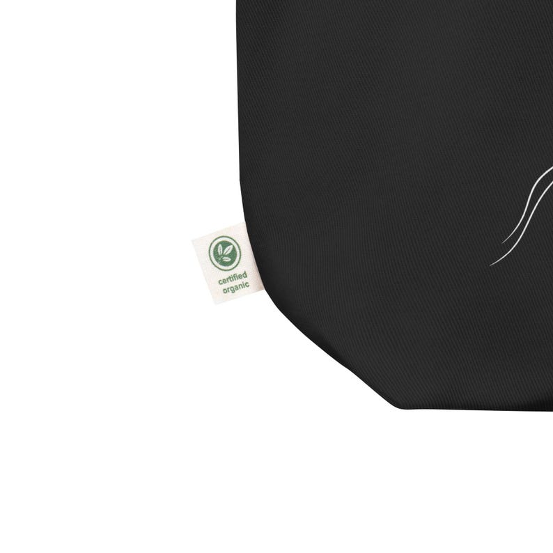 May include: A black fabric tote bag featuring a white line drawing of a plant stem with leaves. A small white rectangular tag is attached to the side, displaying a green circular logo with the text "certified organic" in black lettering.