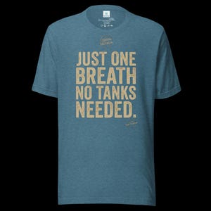 May include: A heathered blue short-sleeve t-shirt featuring the beige text "JUST ONE BREATH NO TANKS NEEDED." printed on the front. The shirt includes a small circular logo above the text and a smaller circular logo near the bottom right corner.