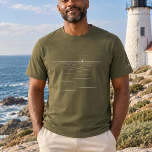Peut inclure: Un t-shirt &agrave; col rond et manches courtes vert olive avec un motif graphique blanc repr&eacute;sentant un petit voilier &agrave; l'horizon sous un ciel &eacute;toil&eacute;, incluant un petit logo blanc sur la manche gauche et un ourlet droit &agrave; la taille.